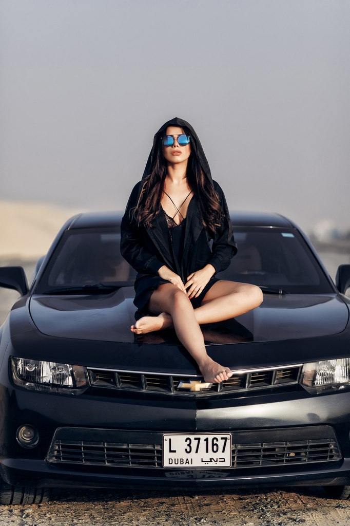 Молодая женщина показывает свою маленькую грудь облокатившись на капот спортивного авто 