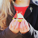 Аватар Блондинистая девушка в в красной водолазке и в черном пальто держит маленькую модель корабля с красным парусом