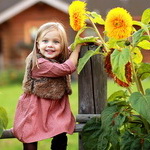 99px.ru аватар Милая, светловолосая, улыбающаяся девочка в меховой жилетке, держащаяся руками за деревянный столбик забора рядом с цветущими желто-оранжевыми георгинами, автор Ирина Сапронова