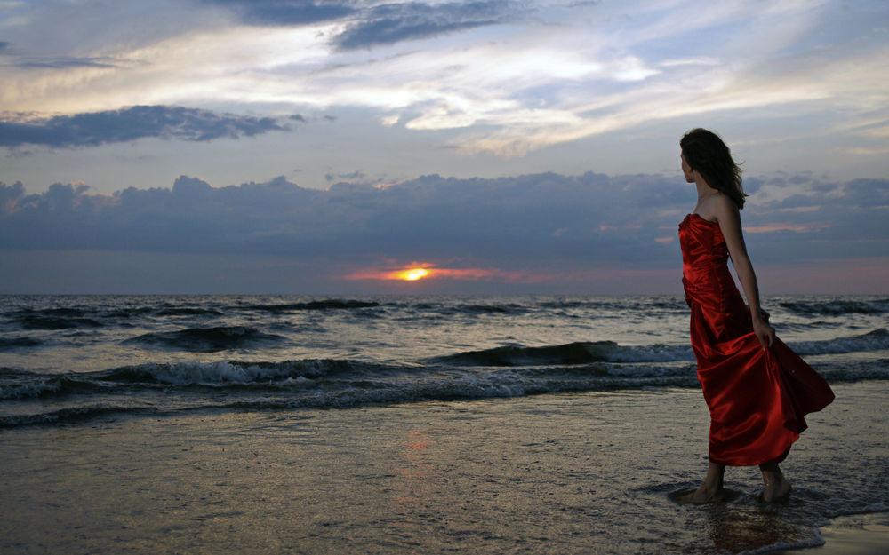 Обои для рабочего стола Девушка в красном платье идет по кромке воды и смотрит на закат