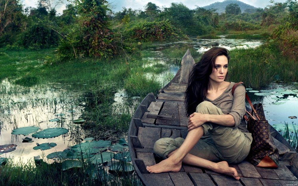 Обои для рабочего стола Анджелина Джолли / Angelina Jolie босая сидит в деревянной лодке с сумкой Луи Витон / Louis Vuitton на плече на фоне болот