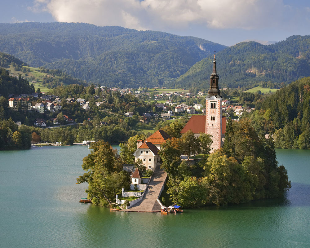 Обои для рабочего стола Маленький остров с церковью посередине озера на фоне не большого города в горах Slovenija / Словении