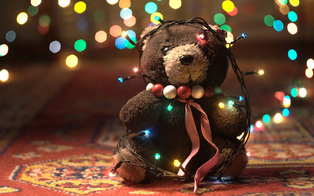 Обои для рабочего стола Плюшевый мишка в бусах, обмотанный новогодней гирляндой с фонариками, сидит на ковре