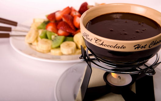 Обои на рабочий стол Чашка горячего шоколада с надписью 'Hot Chocolate' подогревается в шоколаднице, рядом нарезанные фрукты
