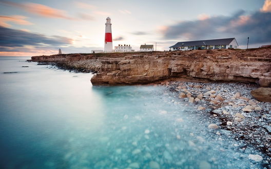 Обои на рабочий стол Маяк стоит на скалистом берегу, омываемом лазурными волнами, рядом с маяком дом смотрителя