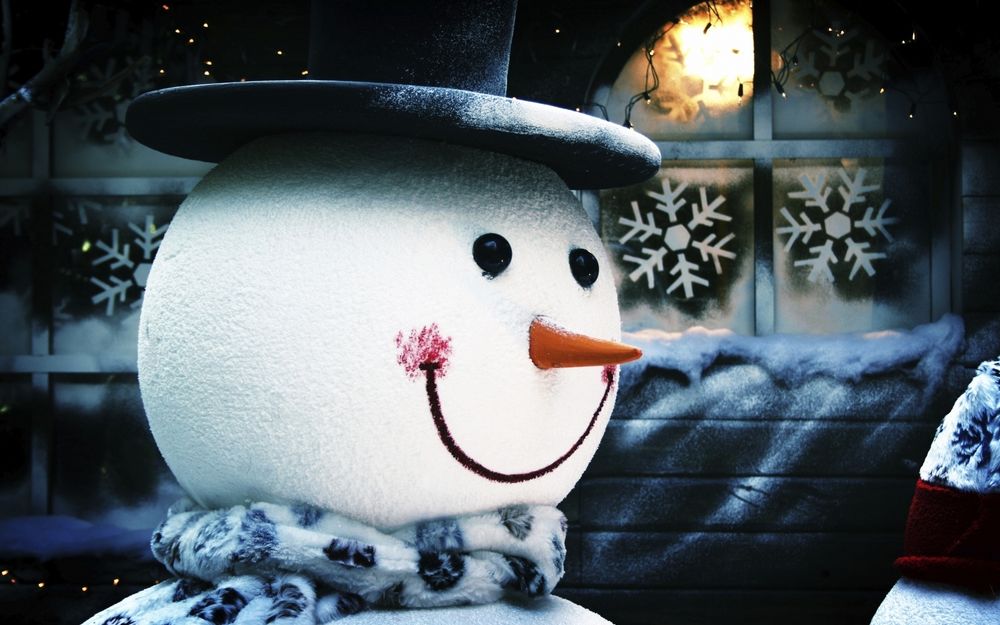 Обои для рабочего стола Весёлый снеговик в шляпе стоит у окна со снежинками