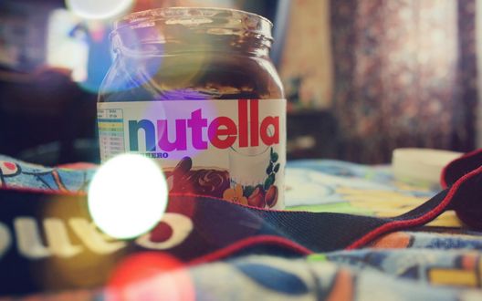 Обои на рабочий стол Банка nutella / нутеллы стоит на кровати, в фотографию попал шнурок от фотоаппарата Canon / Кенон