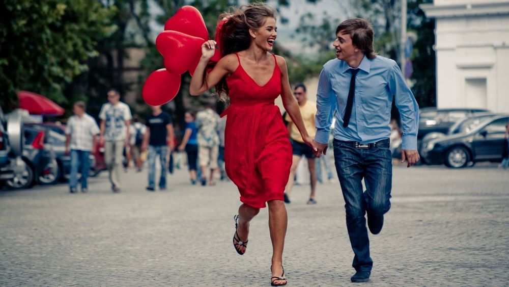 Обои для рабочего стола Девушка держит в руке красные шары в виде сердец и бежит по площади с парнем