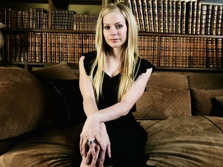 Обои на рабочий стол Аvril Lavigne /  Аврил Лавин в черном платье сидит на фоне многочисленных книг