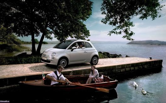 Обои на рабочий стол Автомобиль ФИАТ 500, Италия / Fiat 500, Italy серебристого цвета с женщиной за рулем, стоит на лодочном пирсе на фоне деревьев  и озера, в котором на лодке с веслами  сидят мужчина и женщина