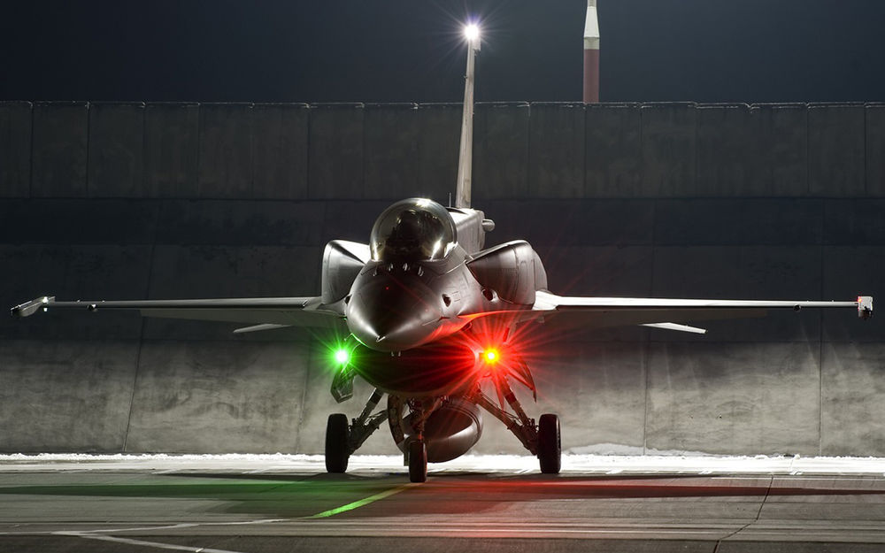 Обои для рабочего стола Американский истребитель F-16 с включенными бортовыми фарами  готовится вырулить на взлетно-посадочную полосу для совершения полета в ночных условиях
