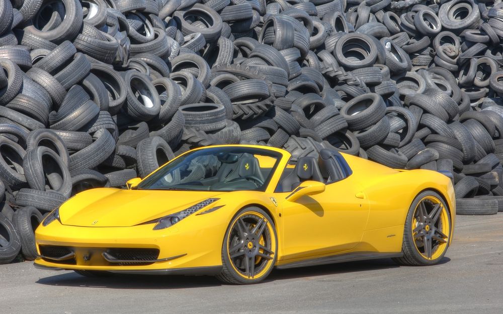 Обои для рабочего стола Легковой автомобиль желтого цвета Ferrari 458 Italia / Феррари 458, суперкар стоит на асфальтированной дороге на фоне большого количества старых автомобильных покрышек