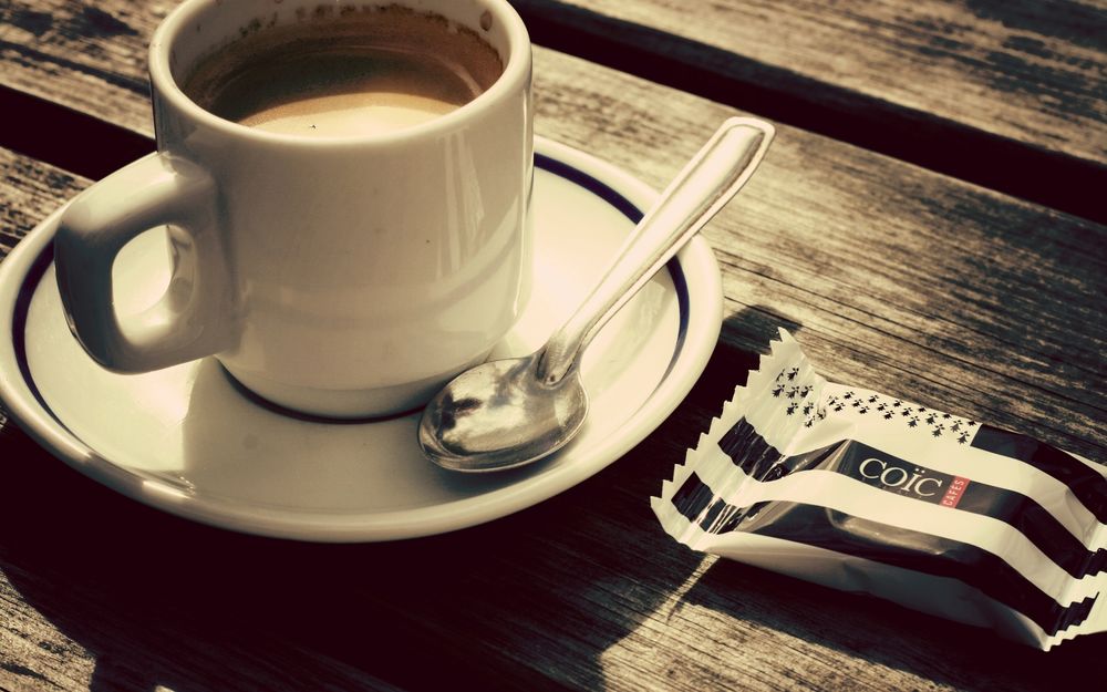 Обои для рабочего стола На деревянном столе стоит белая чашка с кофе, блюдцем и чайной ложкой и рядом находится конфета в обертке (coic cafes) 