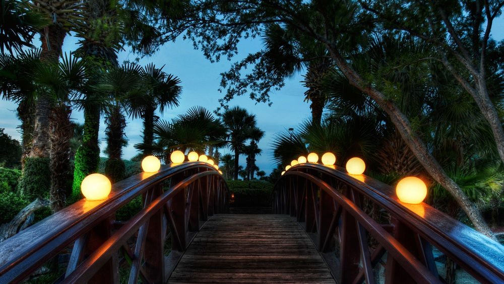 Обои для рабочего стола Деревянный мостик с горящими по бокам фонарями и зелеными пальмами, приглашающий на прогулку по ночному вечернему городу