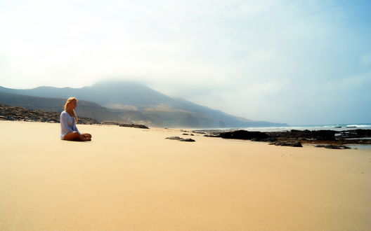 Обои на рабочий стол Блондинка сидит на пляже скрестив ноги на фоне горы