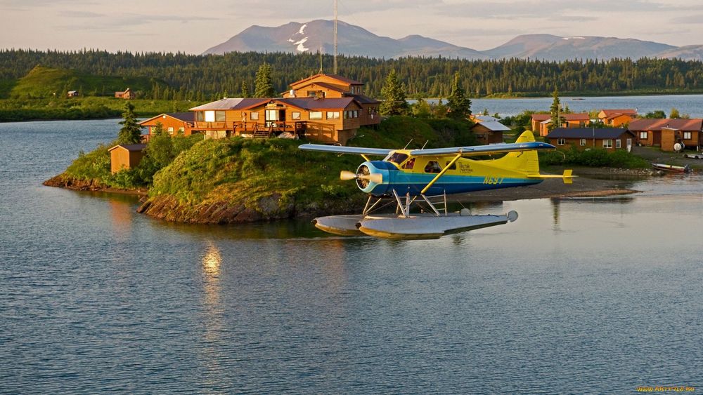 Обои для рабочего стола Самолет-амфибия пришвартовался на водной поверхности озера возле острова с небольшими домиками на полуострове Аляска, США /Alaska, USA на фоне горного массива