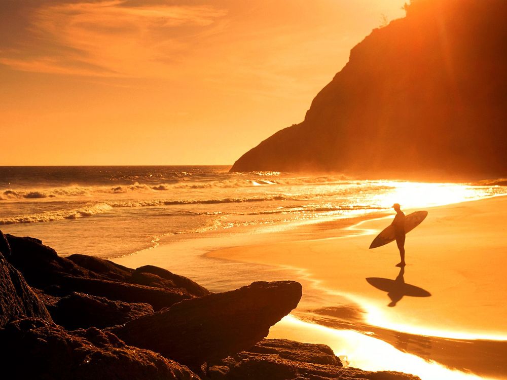 Обои для рабочего стола Одинокий серфингист стоит на песчаном берегу моря, любуясь ярким закатом солнца на фоне горных массивов