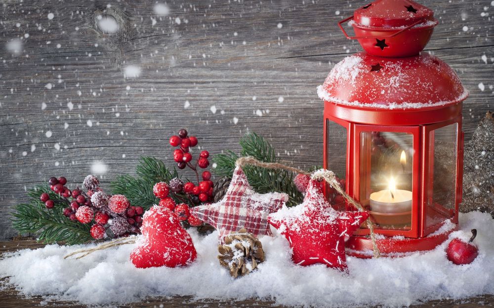 Обои для рабочего стола Красный фонарь с горящей в нем свечой и стоящий на снегу в окружении новогодней праздничной мишуры,  состоящей из:еловых веток, ягод, яблока, шишки и матерчатых подушечек  в виде звездочек и сердечек