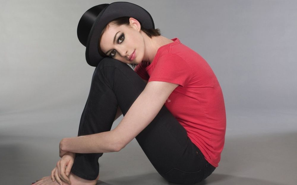 Обои для рабочего стола Американская актриса Энн Хэтэуэй / Anne Hathaway в черных брюках, розовой футболке и шлпе сидит на сером фоне