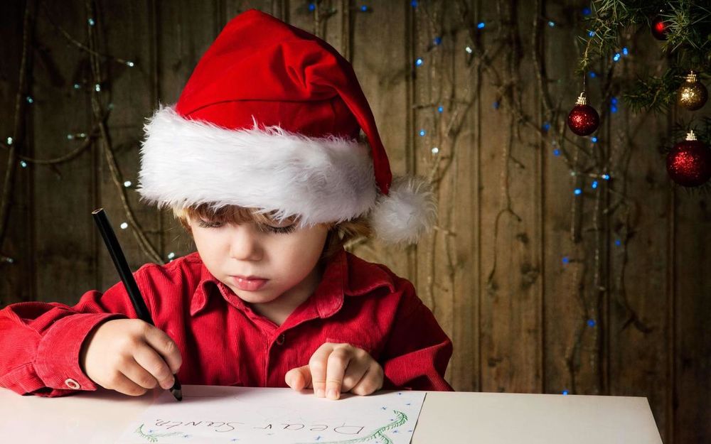 Обои для рабочего стола Маленький мальчик в красной рубашке и новогодней шапочке сидит за столом и пишет письмо Санта Клаусу (Dear Santa)