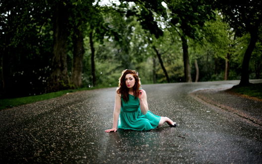 Обои на рабочий стол Странная девушка в зеленом платье сидит на дороге, мокрой после прошедшего дождя