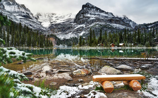 Обои на рабочий стол Чудесное горное образование с небольшим озером с зеленоватой водой, домиками для туристов, на переднем плане деревянная скамейка
