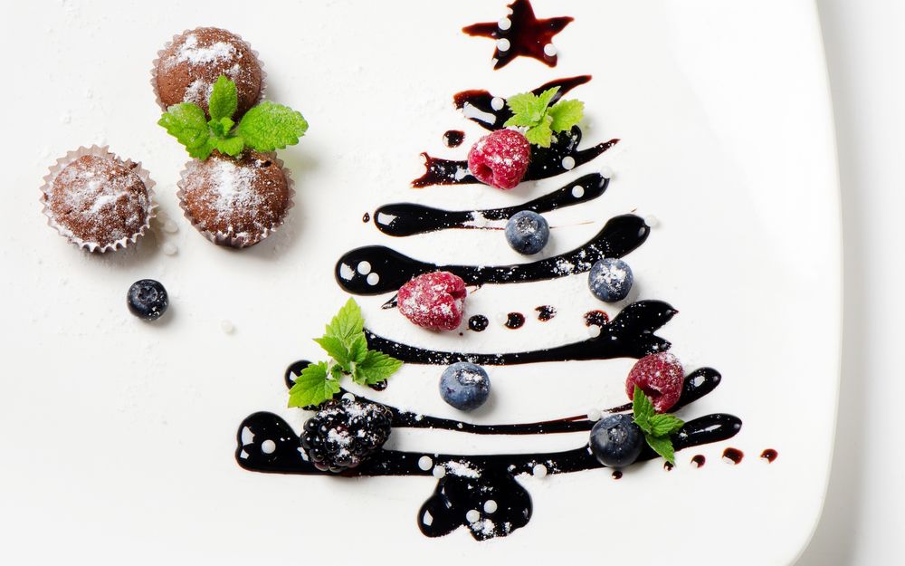 Обои для рабочего стола Ёлка сделанная шоколадной глазурью на тарелке, украшенная маленькими ягодами в виде новогодних игрушек, рядом стоят кексы