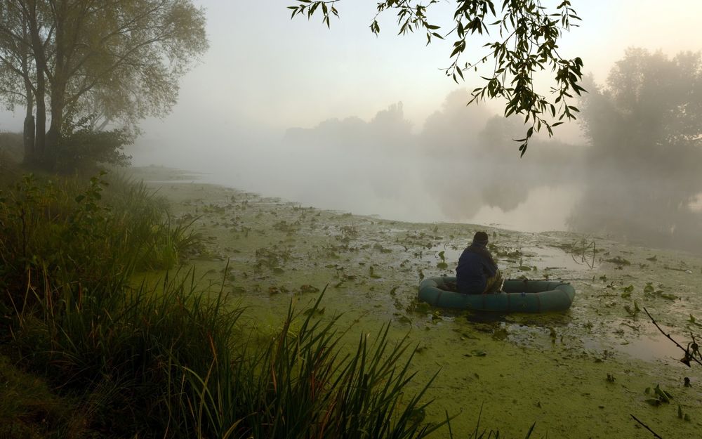 Обои для рабочего стола Рыбак с лежащей удочкой, находящийся в резиновой лодке среди зеленой тины и осоки на озере, покрытом утренним густым туманом