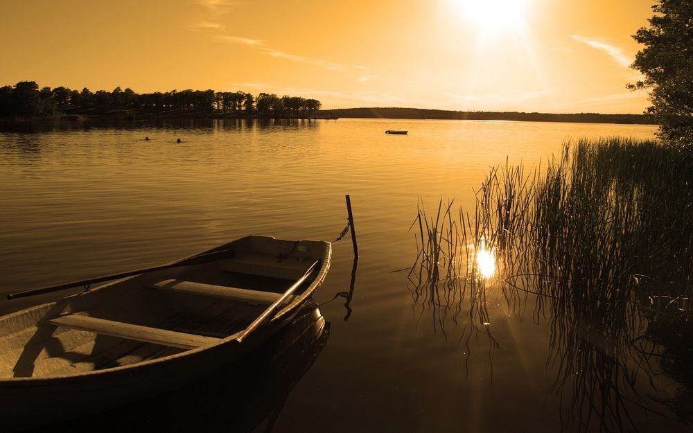 Обои для рабочего стола Рыбацкая лодка, привязанная к торчащей из воды палки возле береговой осоки озера на фоне вечернего неба с заходящим солнцем