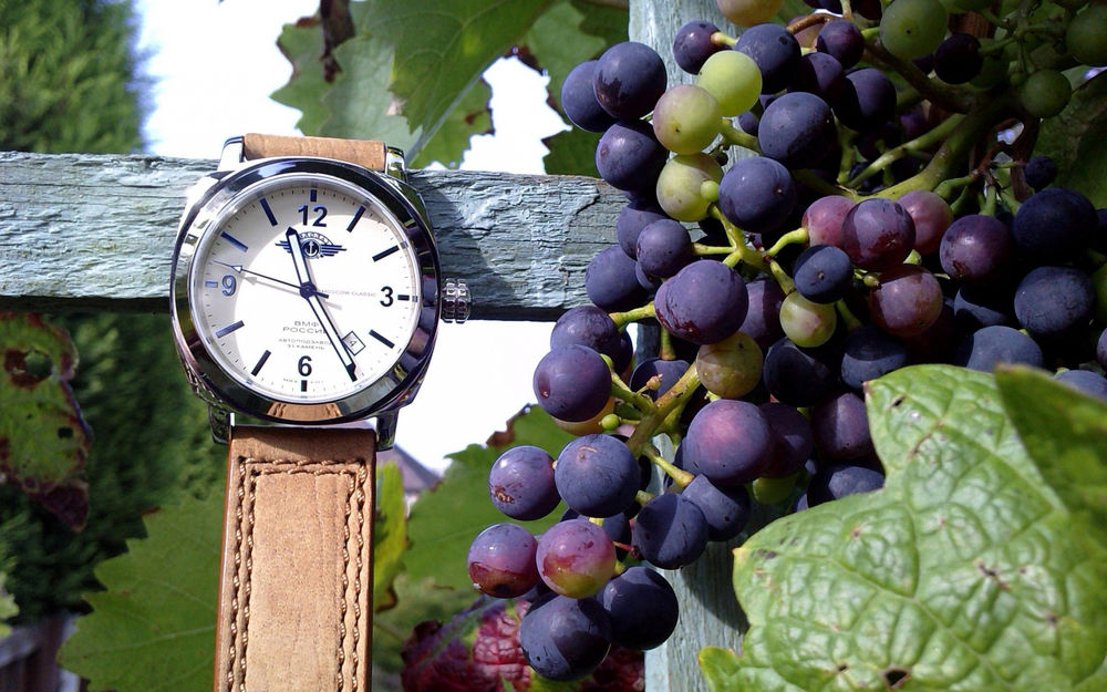 Обои для рабочего стола На деревянной доске висят наручные часы с кожаным ремешком, рядом гроздья винограда на лозе