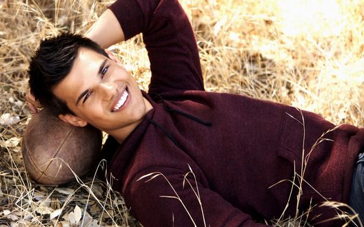 Обои на рабочий стол Американский актер Тейлор Лотнер / Taylor Lautner лежит в траве, подложив под голову мяч для американского футбола, и улыбается