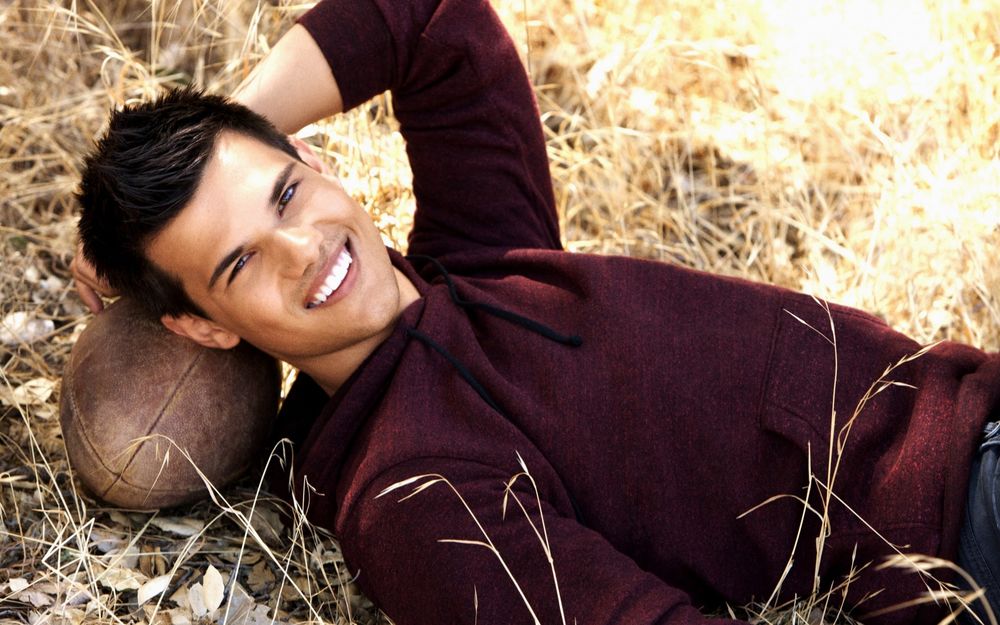 Обои для рабочего стола Американский актер Тейлор Лотнер / Taylor Lautner лежит в траве, подложив под голову мяч для американского футбола, и улыбается