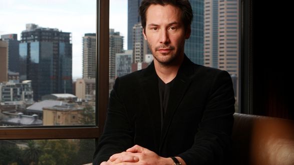 Обои на рабочий стол Киану Чарльз Ривз / Keanu Charles Reeves в черном пиджаке сидит на на фоне города
