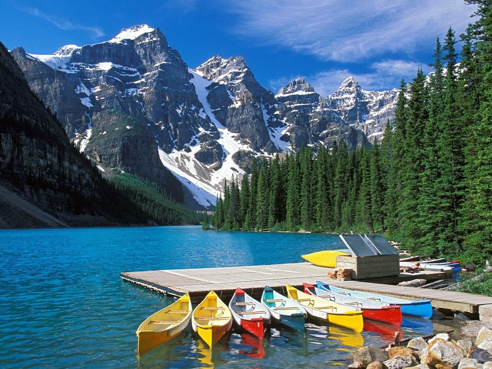 Обои для рабочего стола Деревянный лодочный причал с разноцветными прогулочными лодками, стоящий на берегу красивого горного озера, мужчина-лодочник загорает в ожидании туристов