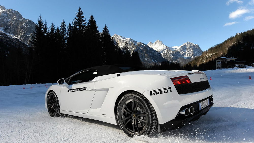 Обои для рабочего стола Белый Lamborghini Gallardo LP 560-4 стоит на снегу, на фоне дома, гор и деревьев