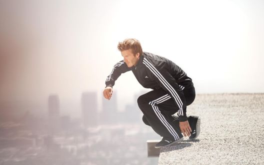 Обои на рабочий стол Дэвид Бекхэм / David Beckham в черных кроссовках и спортивном костюме в белую полоску от Адидас / Adidas собирается прыгнуть с крыши многоэтажного дома