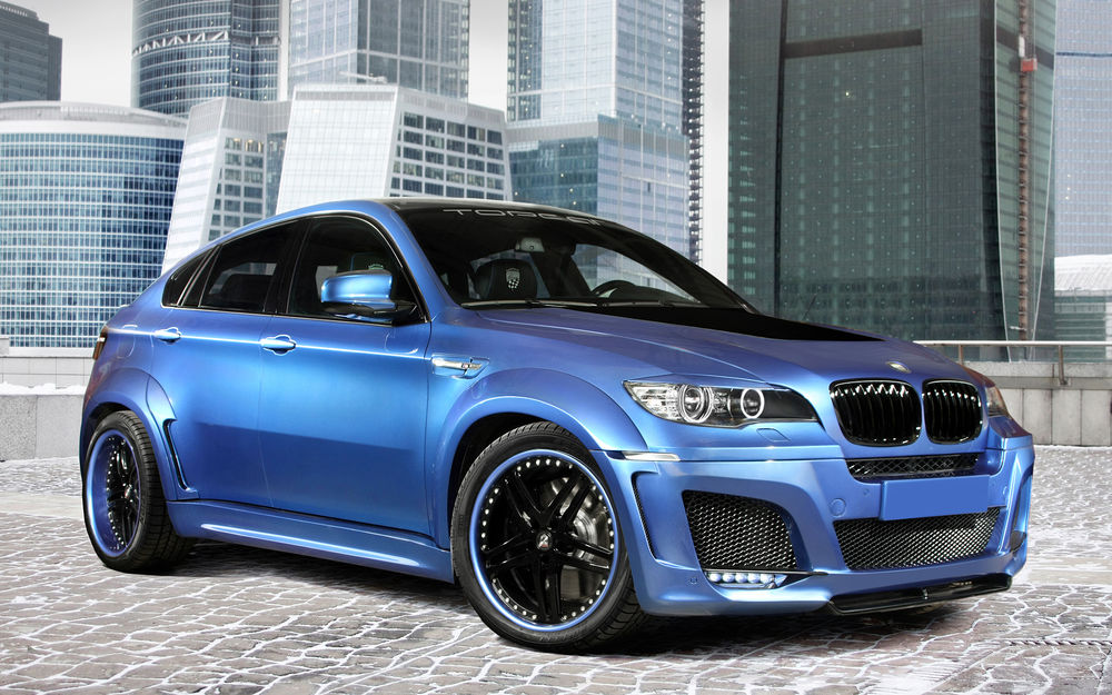 Обои для рабочего стола Автомобиль BMW X6 голубого цвета стоит на фоне небоскребов мегаполиса