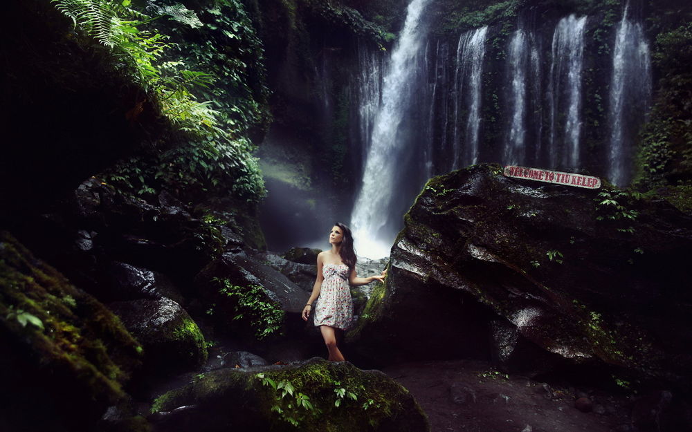 Обои для рабочего стола Девушка шатенка в платье стоит на фоне знаменитого водопада Tiu Kelep, расположенного на острове Ломбок, Индонезия / Lombok, Indonesia, на камне надпись WELCOME TO TIU KELEP / Добро пожаловать на водопад Tiu Kelep
