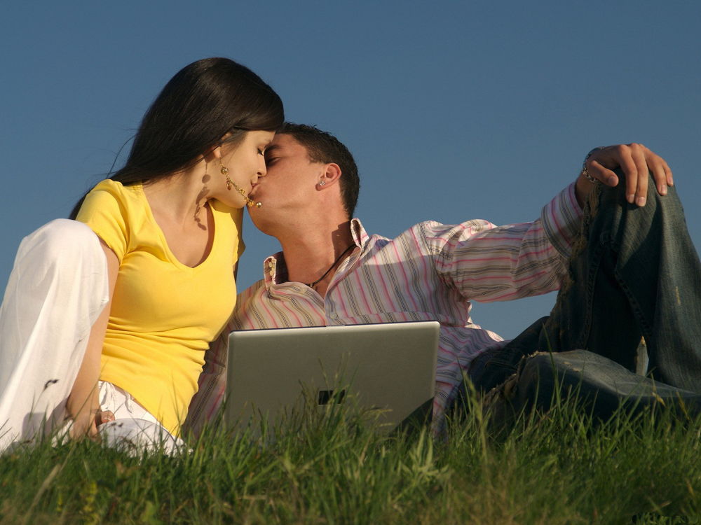 Обои для рабочего стола Целующиеся парень и девушка, сидящие на траве возле открытого ноутбука на фоне голубого неба