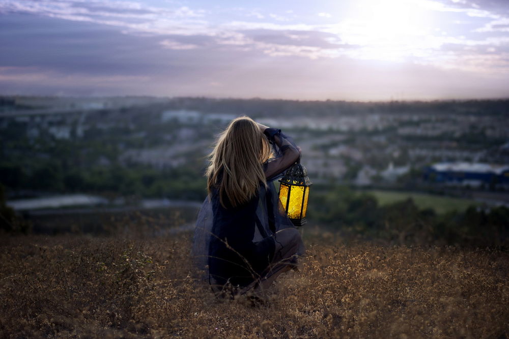 Обои для рабочего стола Девушка с фонарем сидит на вершине холма, а внизу город