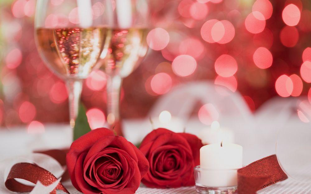 Обои для рабочего стола Две красные розы лежат на столе рядом со свечой, на заднем плане стоят да бокала