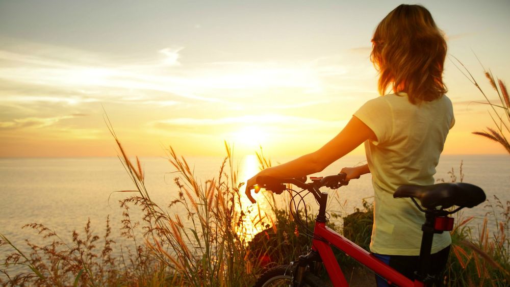 Обои для рабочего стола Девушка стоит с велосипедом в высокой траве и смотрит на закат над морем