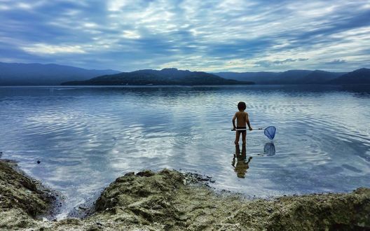 Обои на рабочий стол Мальчик, стоящий в береговой воде озера, держащий в руках садок для рыбы на фоне пасмурного неба