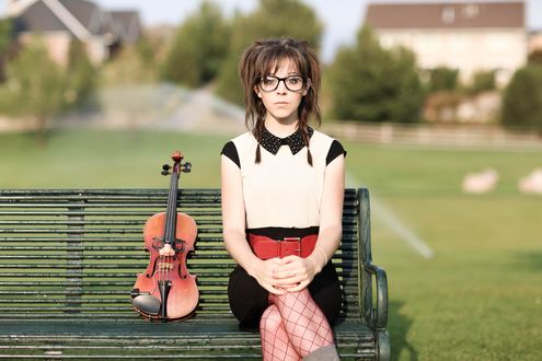 Обои на рабочий стол Линдси Стирлинг / Lindsey Stirling сидит на скамейке в красных чулках в крупную сетку, в очках, рядом со скрипкой на фоне загородных домов