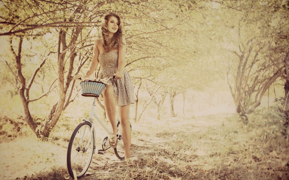 Обои для рабочего стола Девушка в легком летнем сарафане едет на велосипеде среди низких деревьев