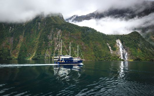 Обои на рабочий стол Синий корабль плывет по фьорду Милфорд Саунд / Milford Sound, мимо водопада Боуэн / Bowen Falls, Новая Зеландия / New Zealand
