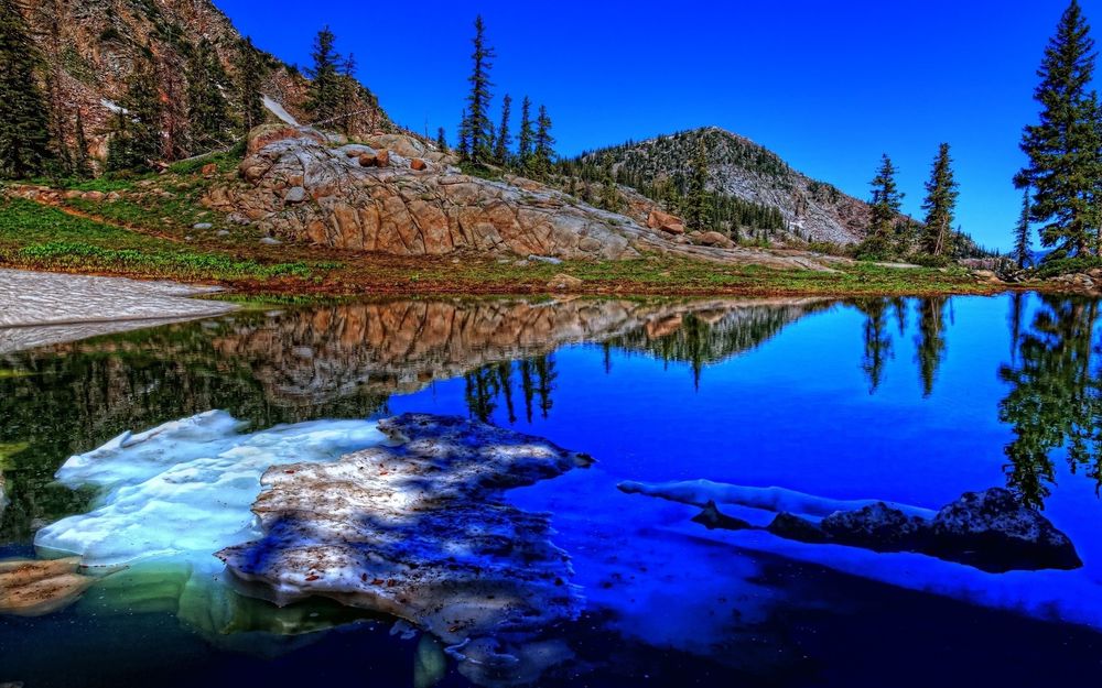 Обои для рабочего стола Берег озера с красивыми невысокими скалами, покрытыми высокими соснами, отражающимися в зеркальной водной глади с плавающими небольшими льдинами на фоне яркого голубого неба