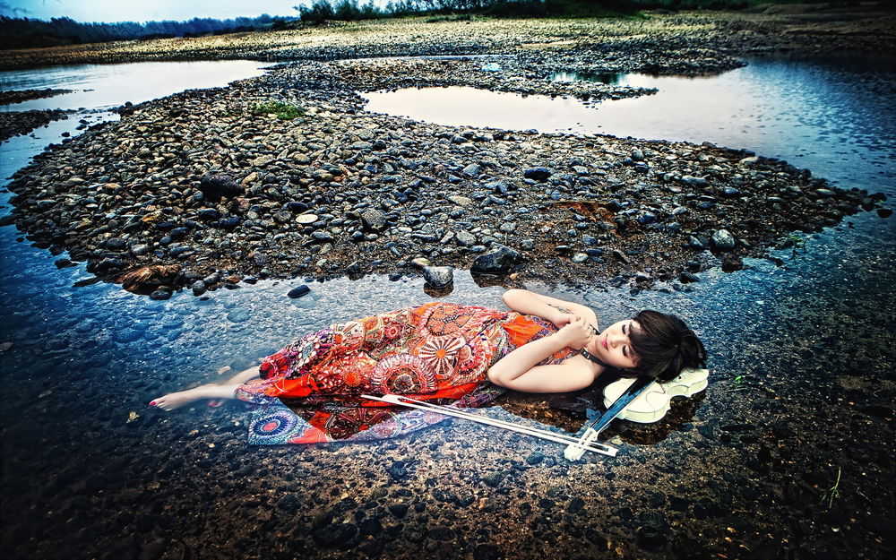 Обои для рабочего стола Девушка восточной внешности лежит в воде у каменистого берега, положив голову на белую скрипку