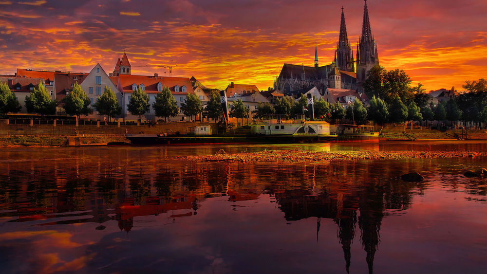 Обои для рабочего стола Вид на вечерний Регенсбург, Германия / Regensburg, Germany, небольшой корабль пришвартован у берега Дуная, над которым возвышаются две башни кафедрального собора св. Петра / Kathedrale St. Peter