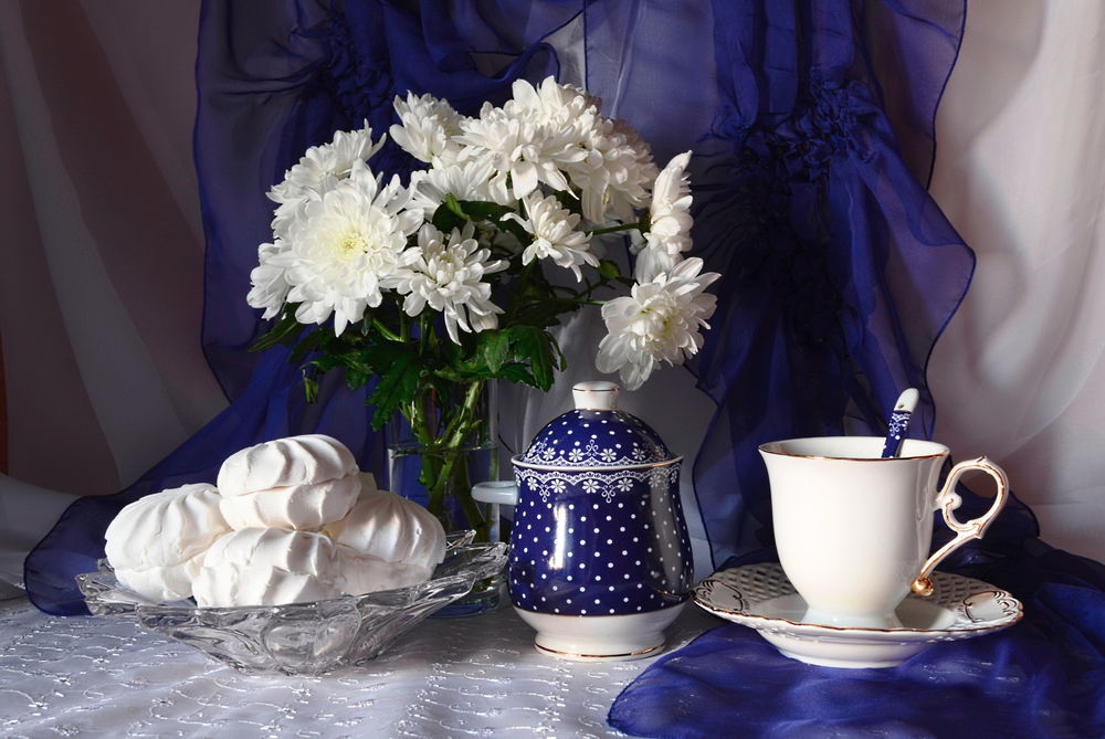 Обои для рабочего стола На столе на белой скатерти на фоне синей драпировки стоит стеклянная ваза с водой в которой находятся нежные, белые цветы хризантемы, в стеклянной чаше лежит белый зефир, рядом стоит сахарница и чашка для чая с блюдечком и ложкой